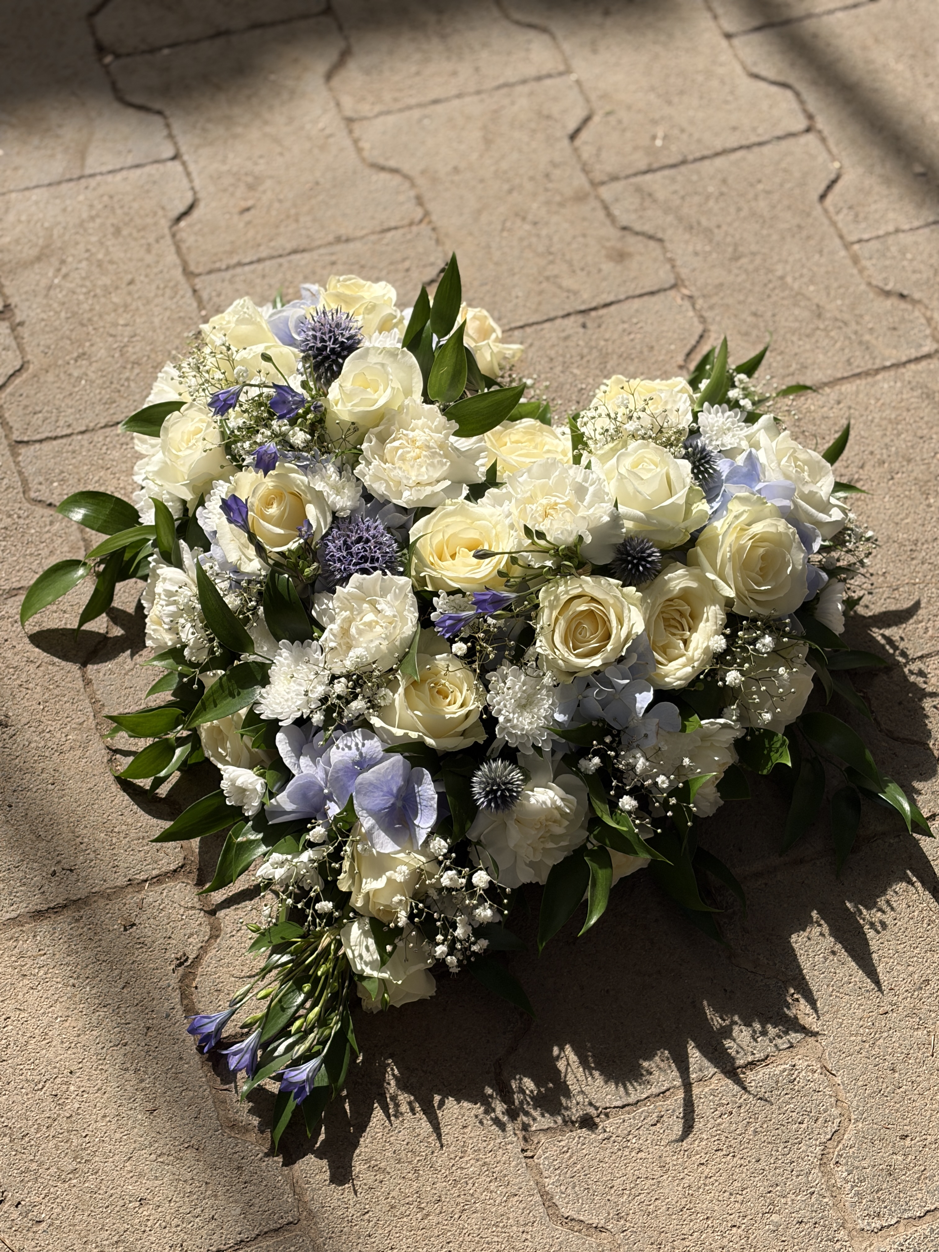 Herzförmiges Trauergesteck mit weißen Rosen, hellblauen Blüten und frischem Grün